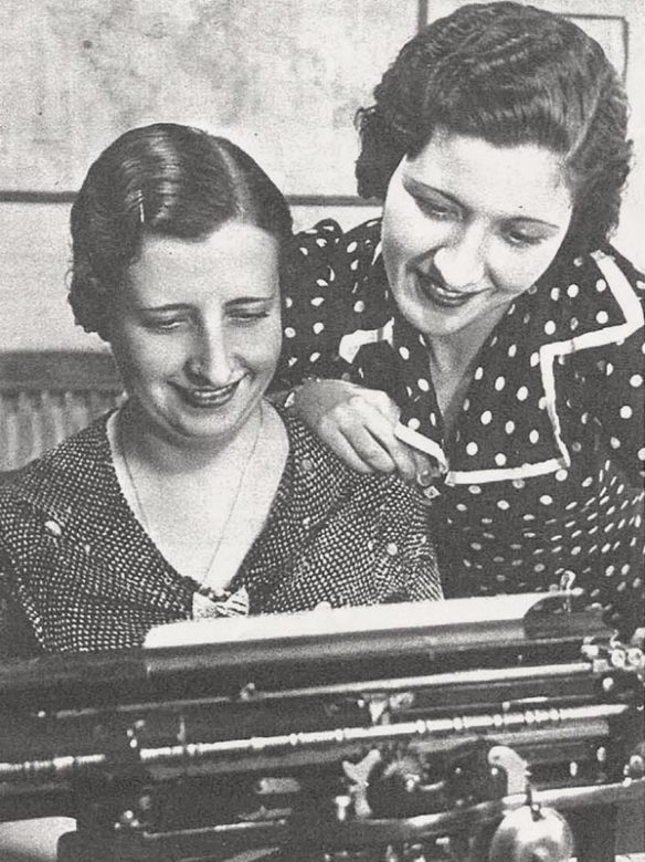 Fotografía original del artículo. Una mujer frente a una máquina de escribir, y otra apoyada en su hombro. Ambas miran el texto y sonríen.